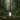 A stacked candle sculpture stands on mossy ground in a forest setting. The design features a cream Buddha head on a dark red base, stacked with pink and dark purple textured rings, topped with a small red apple-shaped candle.