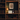 A warm, cozy interior scene showing a wooden bookshelf filled with books, featuring a lit table lamp, a white laptop labeled 'Dulann', a small decorative figurine, and a framed abstract print on a dark wall.