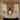 A decorative stacked candle holder or sculpture sits on a white marble mantel in a bright interior room with tall windows and cream curtains. The object has a white sculptural head base, dark purple stacked rings, pink textured bands, a red candle vessel, and a lit candle.