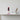 A styled interior scene shows a decorative stacked sculpture on a white pedestal with a pink base, next to a white card reading 'Dulenn', and a small white vase with dried pampas grass on a marble console table against a textured white wall.